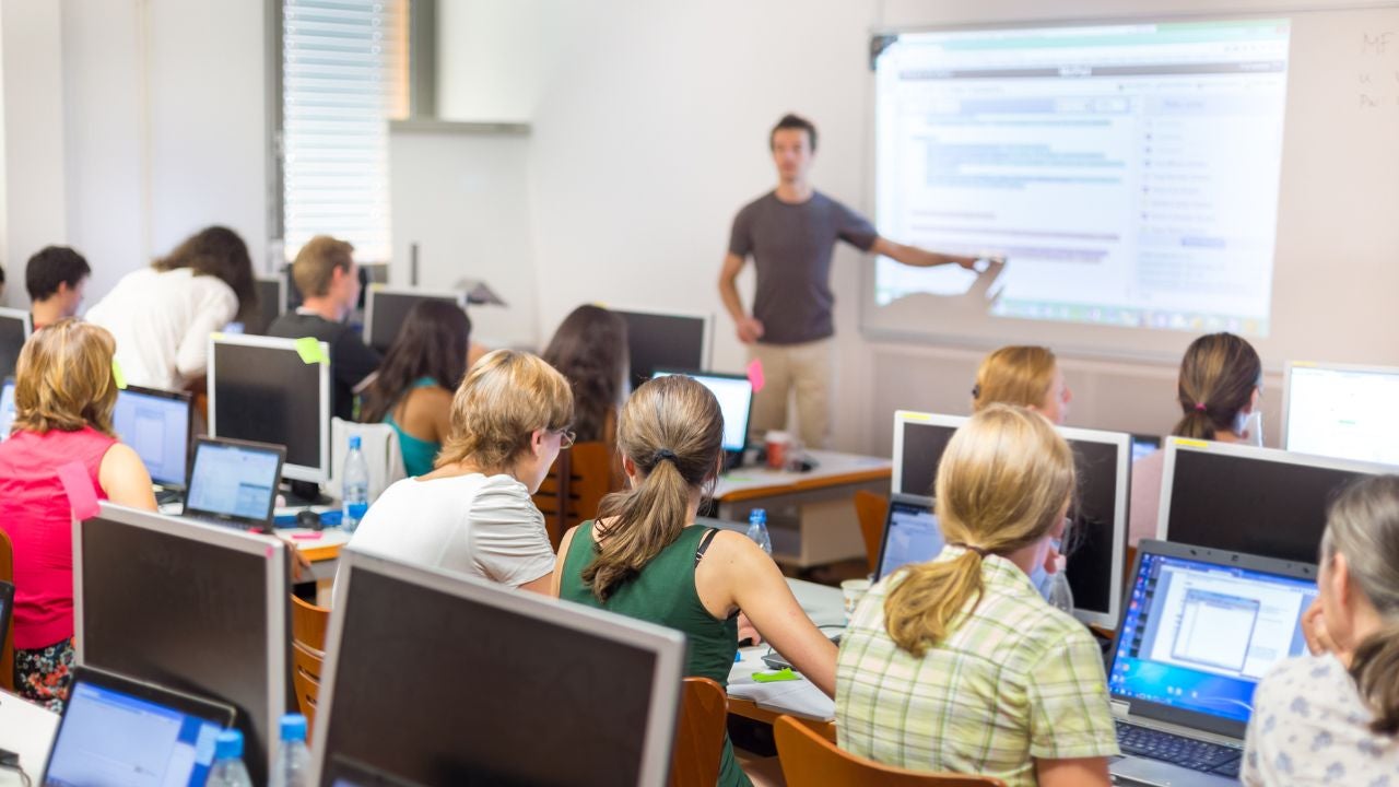 A teacher presents at the front of a classroom with students seated at tables with computers.