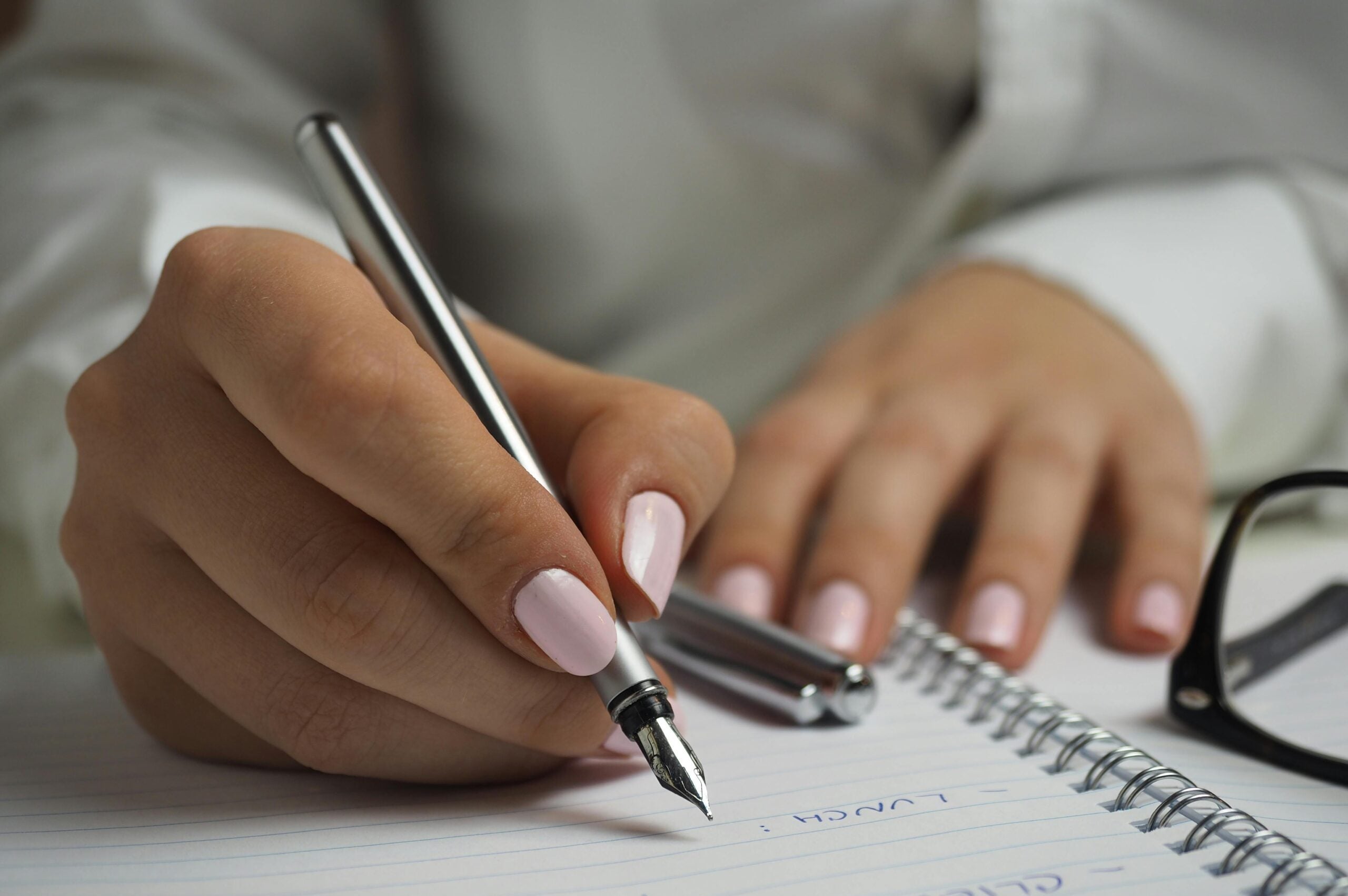 Hands writing notes with a pen, glasses nearby.