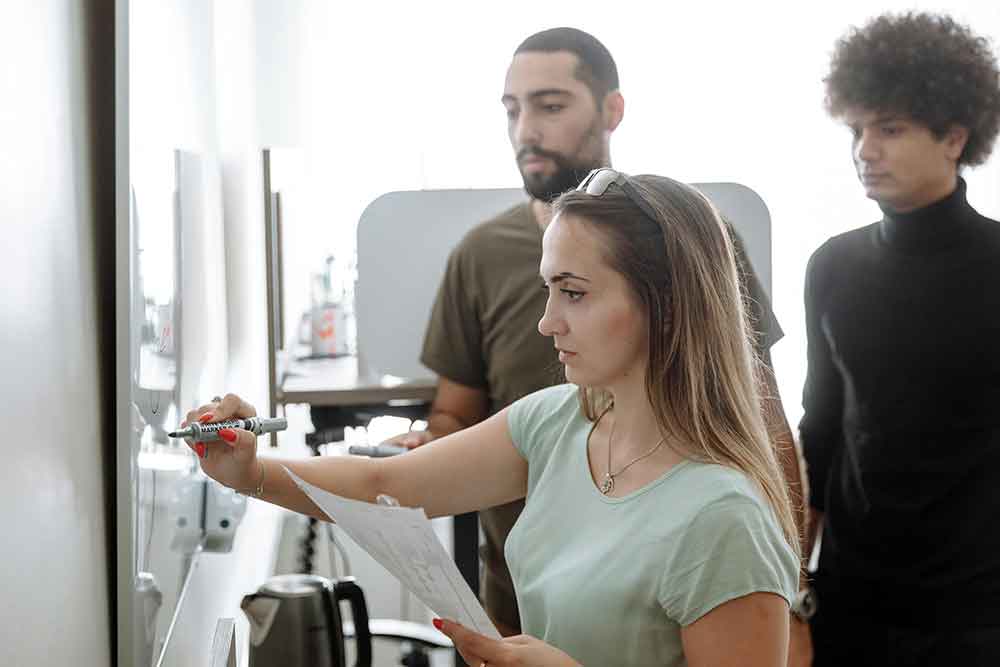 Female instructor writing on white board with two male students watching.