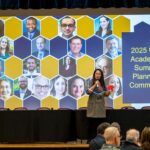 URI Provost speaking on a stage with a background image showcasing many individual headshots in a honeycomb graphic.