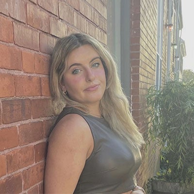 URI student with long blond hair leaning against a brick wall