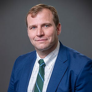 Image of a man, URI DBA student, wearing a blue suit with a green striped tie smiling.