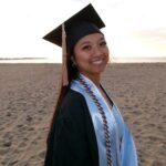 URI college of business student wearing a graduation gown and cap, with the beach as a background.