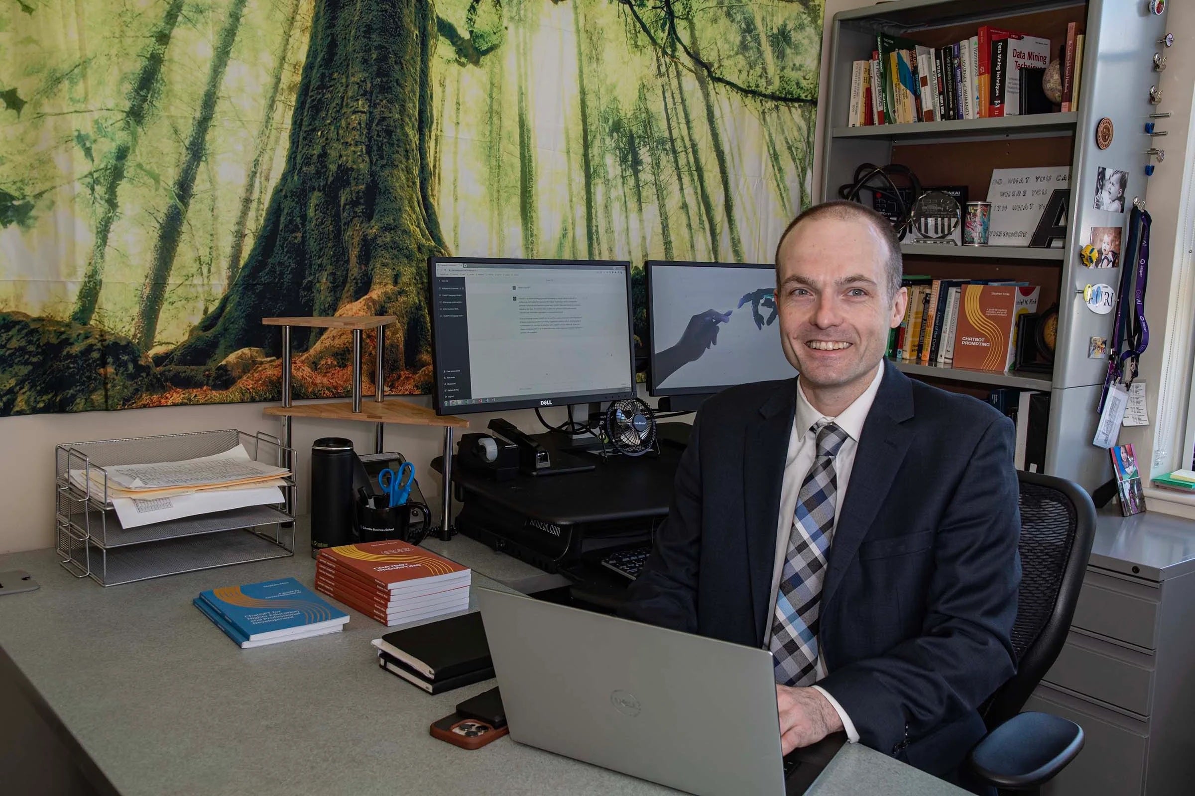 URI professor sitting at a desk surrounded by computers and book shelves