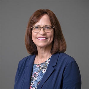 woman with glasses smiling wearing a blue coat and patterned shirt