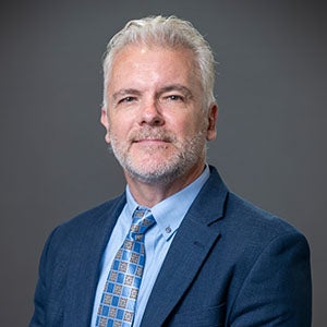 A professional portrait of a man, URI DBA student, with grey hair and a beard, wearing a blue suit, light blue shirt, and patterned tie, set against a plain dark background