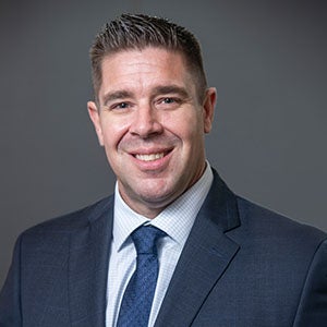 image of a man, URI DBA student, in a dark blue suit with a blue tie, smiling with a muted gray background