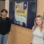 Two URI College of Business students standing against a wall that has an image on it