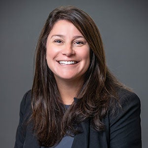 A professional portrait of a woman, URI DBA studetn, with long brown hair, smiling, wearing a dark blazer, set against a plain dark background.