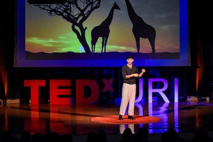 TEDxURI event with a person on the stage and giraffes on the screen behind him