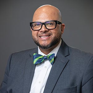 A professional portrait of a man, URI DBA student, in glasses, wearing a grey suit, white shirt, and colorful bow tie, set against a plain dark background.