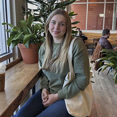 URI student sitting on a stool with plants in the background and coffee on the table