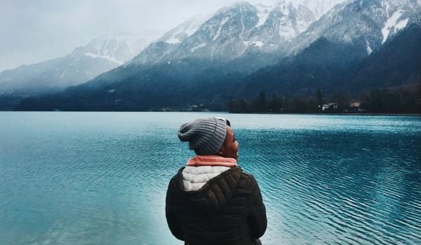 woman sits in front of a lake and snow-covered mountains