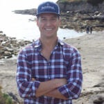David Bourbeau standing with his arms crossed on the beach wearing a plaid shirt and a baseball hat