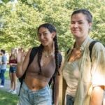 image of 2 students smiling looking at the camera while in line at the farmers market with trees in the background and a line of students gathered in the background