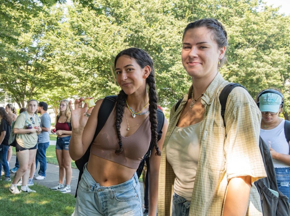 image of 2 students smiling looking at the camera while in line at the farmers market with trees in the background and a line of students gathered in the background