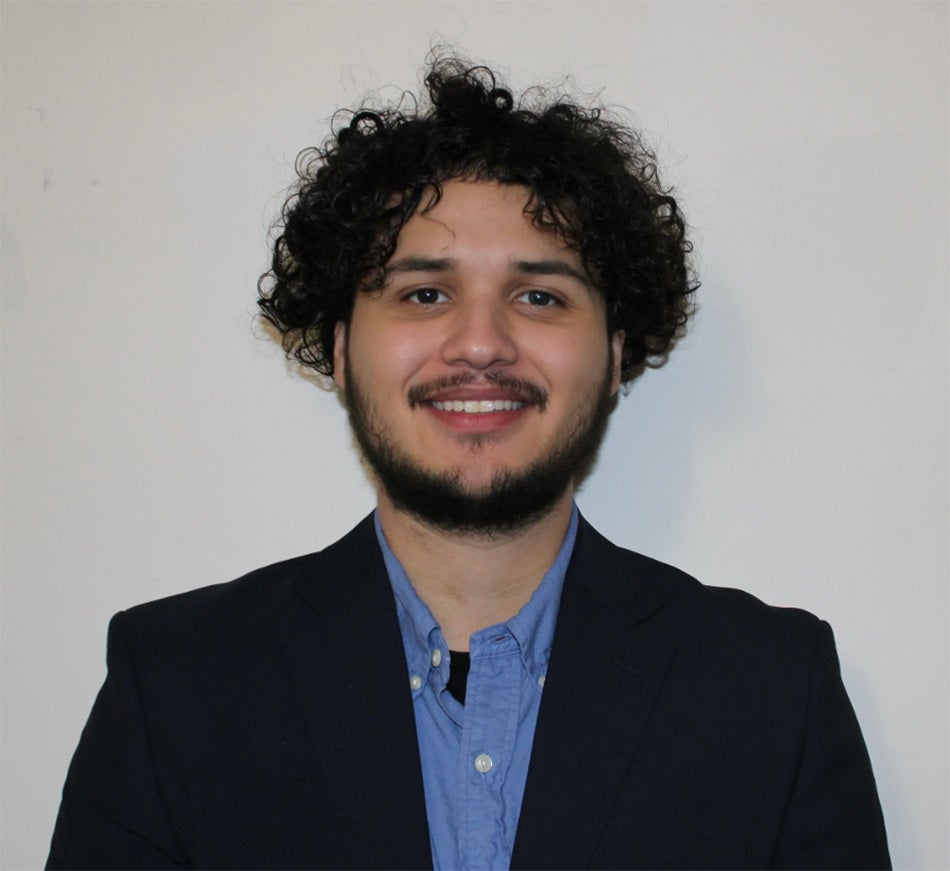 image of Alberto Paz standing in front of a gray background. He is wearing a blue collared shirt with a dark navy blazer. He is smiling at the camera.