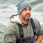 Perry Raso ’02, M.S. ’06, founder and owner of Matunuck Oyster Farm, stands waist-deep in Potter Pond, in South Kingstown, R.I. standing in a cove holding oysters from an oyster farm