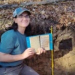 Rachel Howard in a soil pit during a wetland ecology field lab (NRS 423), holding a book used to describe soil color by hue, value, and chroma.