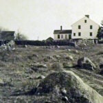 A black and white image of a white colonial-style home next to a stone wall and rocky yard