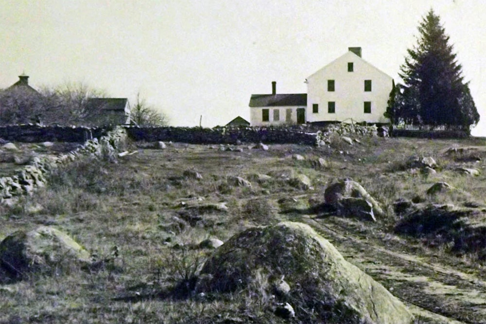 A black and white image of a white colonial-style home next to a stone wall and rocky yard