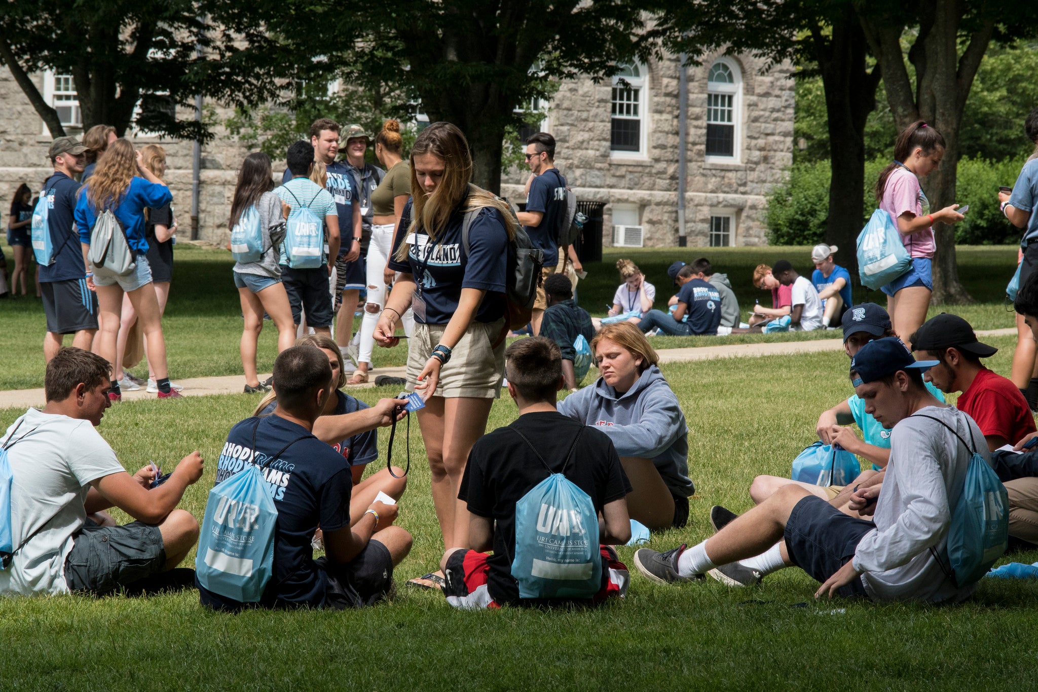 Photograph of students or people standing or walking on a university campus lawn in front of academic buildings, with trees and blue sky visible.