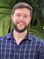 Tyler Delaney, Postdoctoral Fellow, wearing a blue shirt standing in front of a tall plant