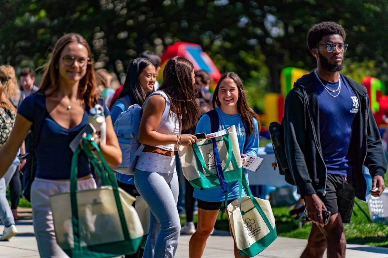 A group of college students walk outdoors on a sunny day, smiling and talking while carrying tote bags, with colorful inflatable activities and other students in the background, suggesting a lively campus event.