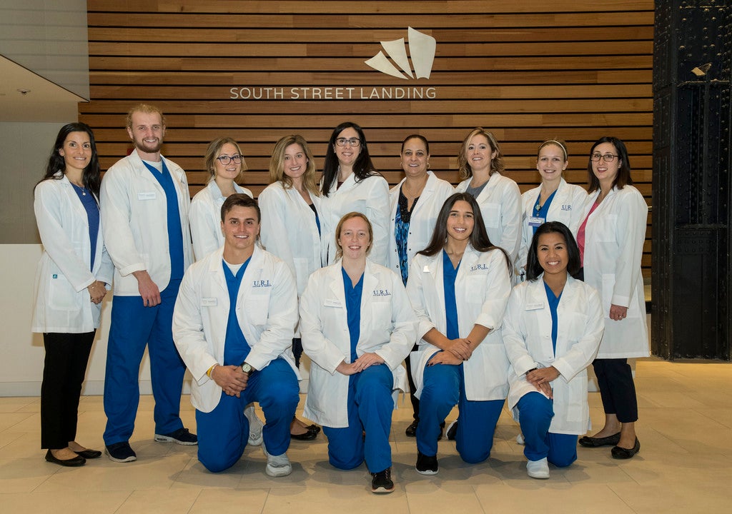 Students in lab coats and blue scrubs posing in front of a South Street Landing sign
