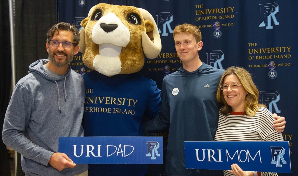 A student stands smiling beside the University of Rhode Island’s Ram mascot, Rhody, in front of a branded URI backdrop, suggesting a welcoming campus environment and school spirit.