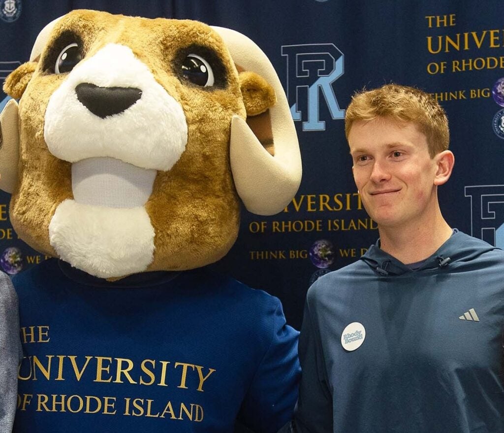 A student stands smiling beside the University of Rhode Island’s Ram mascot, Rhody, in front of a branded URI backdrop, suggesting a welcoming campus environment and school spirit.