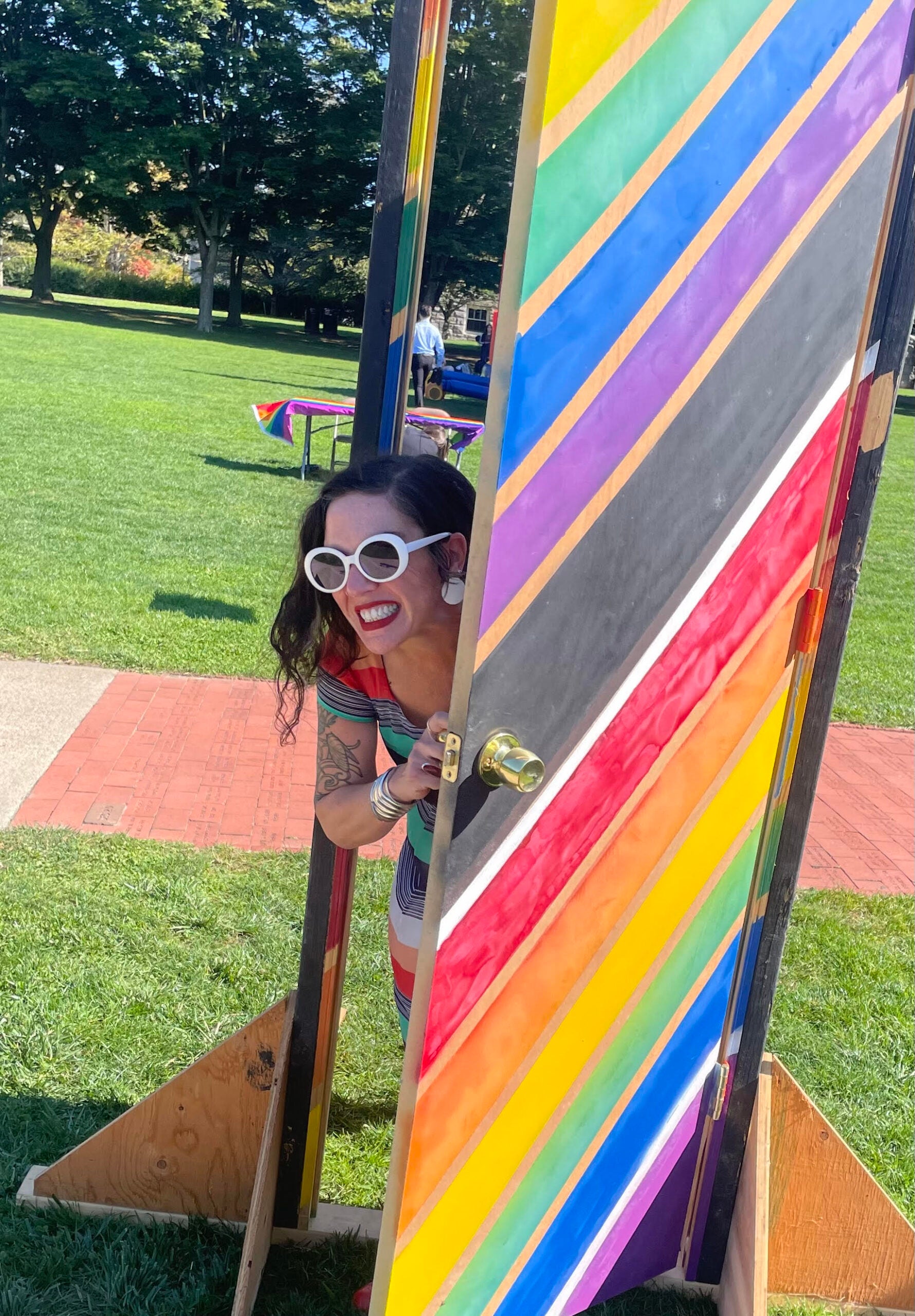 A smiling white person with dark hair, wearing sunglasses peeks out from behind a rainbow door