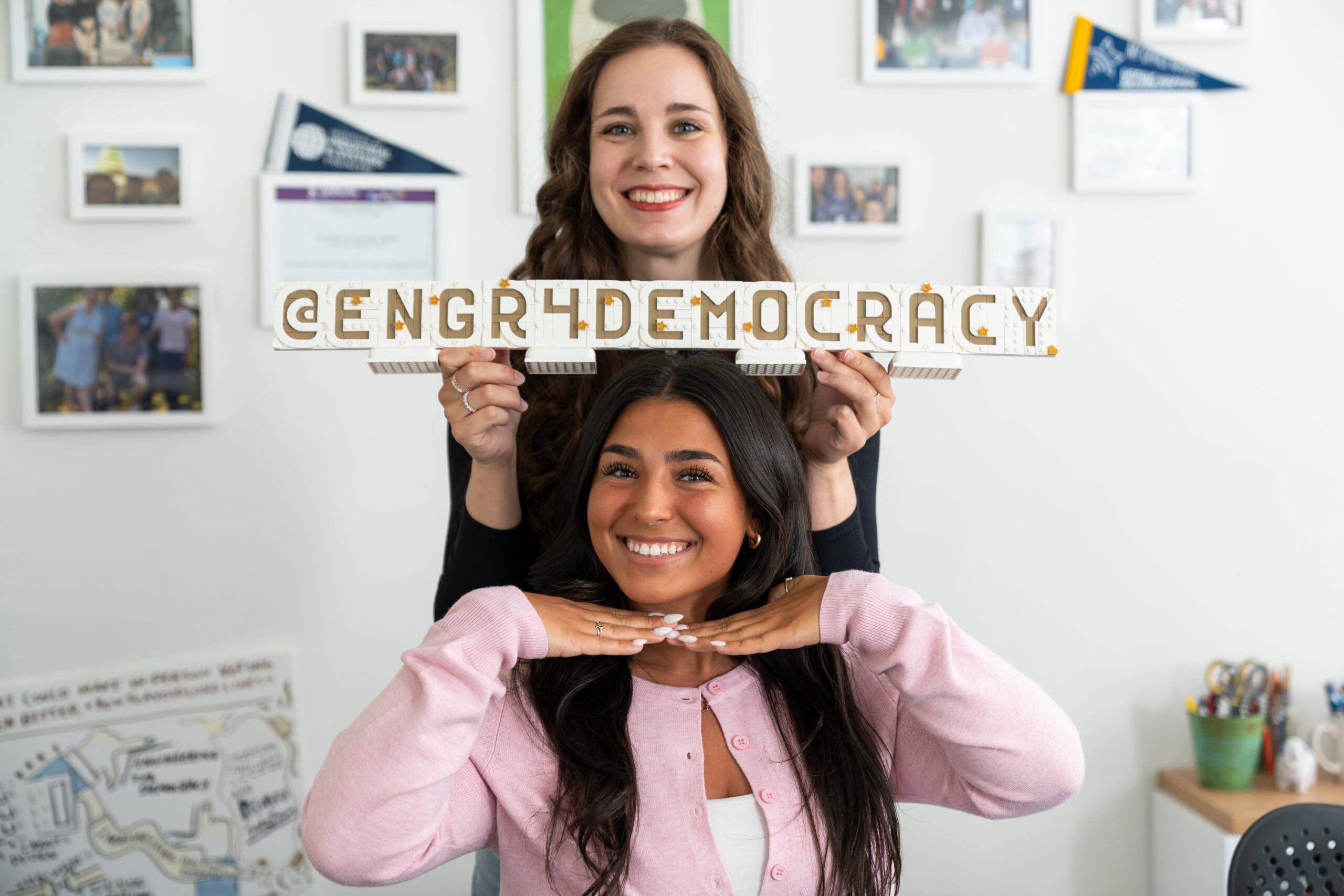 Two smiling women in a bright office; one holds a white and gold LEGO sign reading “@ENGR4DEMOCRACY” above the other’s head.