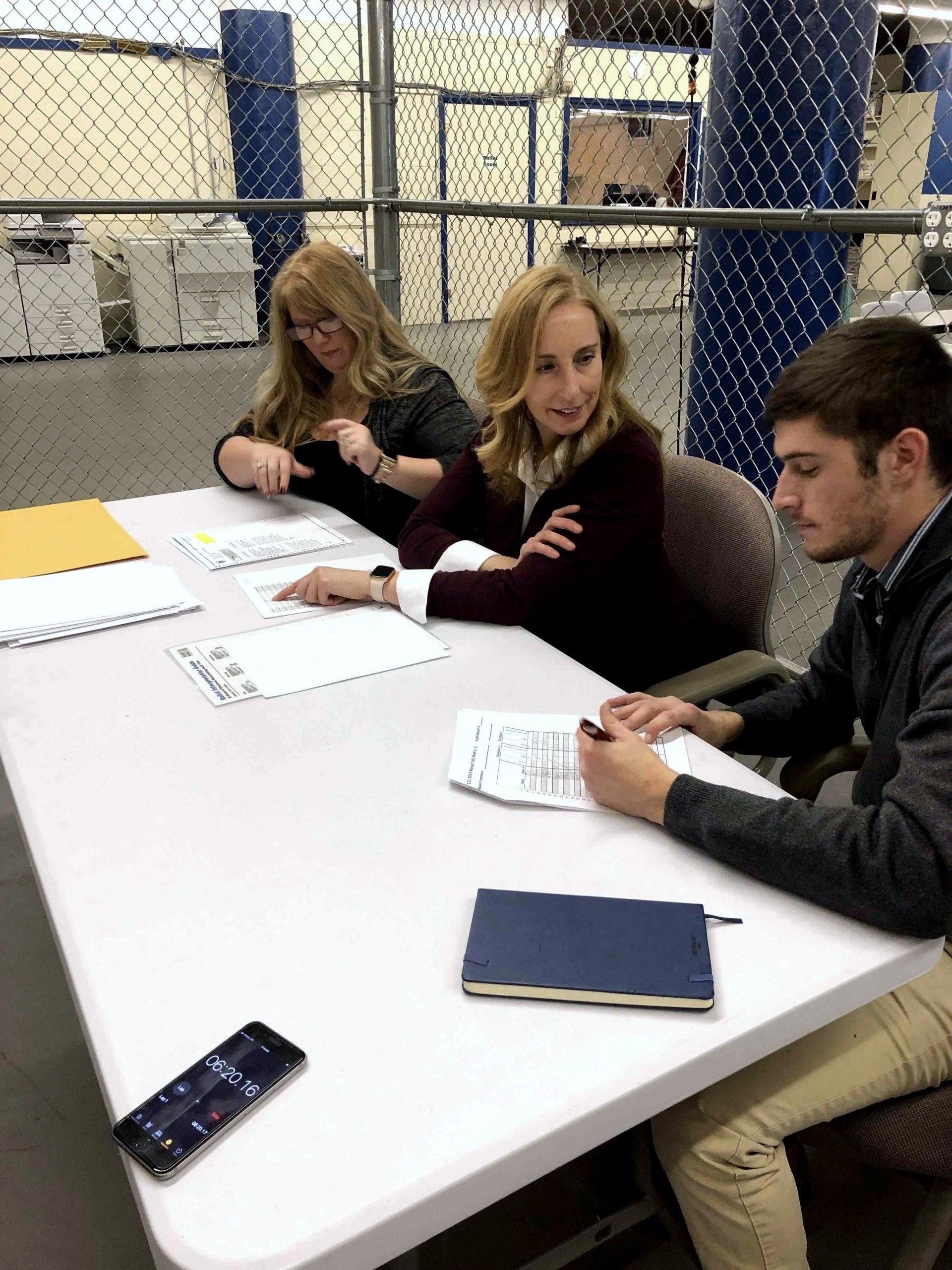 Three election workers seated at a table reviewing ballots and tally sheets in a warehouse.