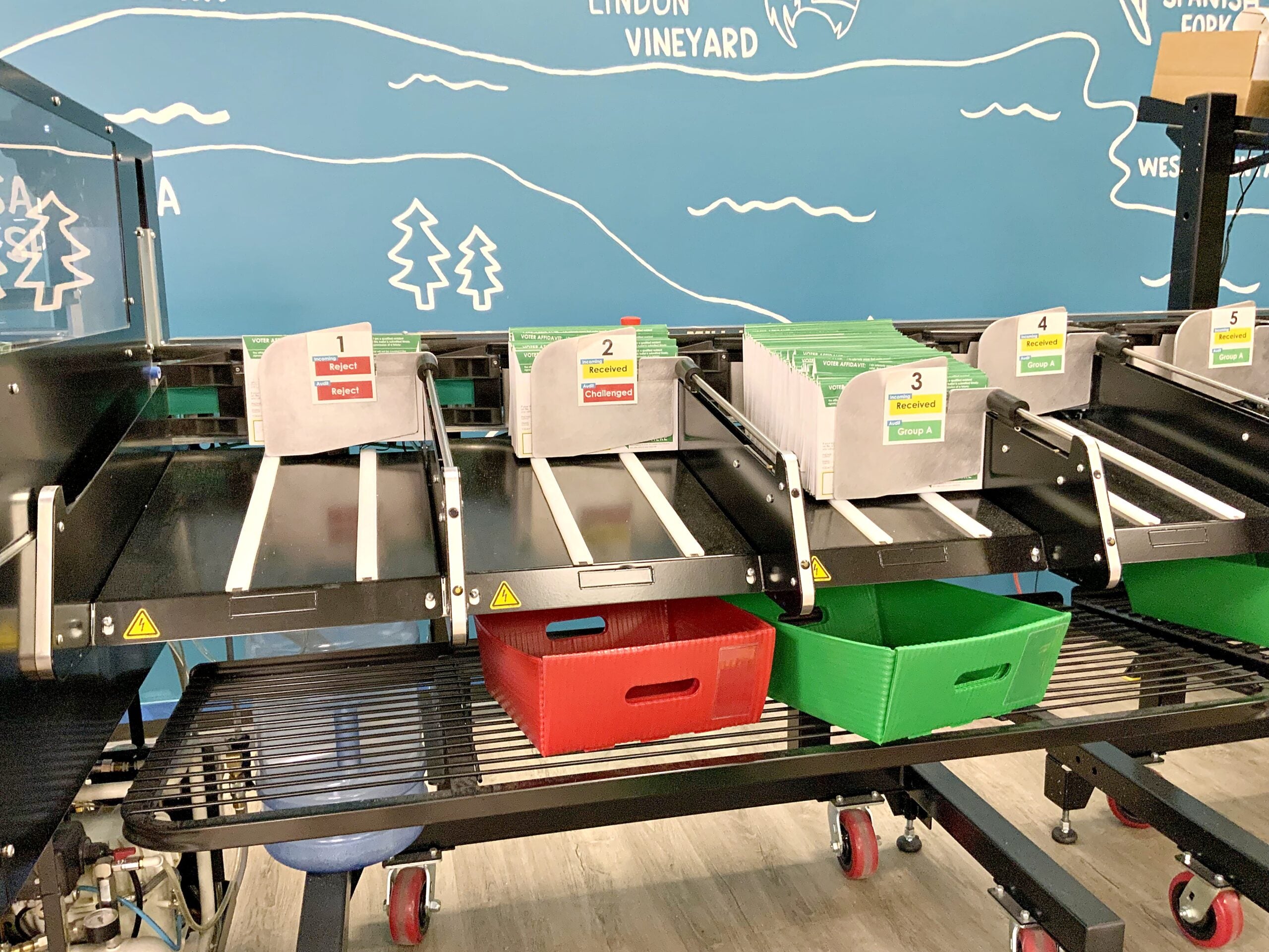 A mail-ballot sorting machine with numbered trays labeled Reject, Received, and Group A, in front of a blue mural