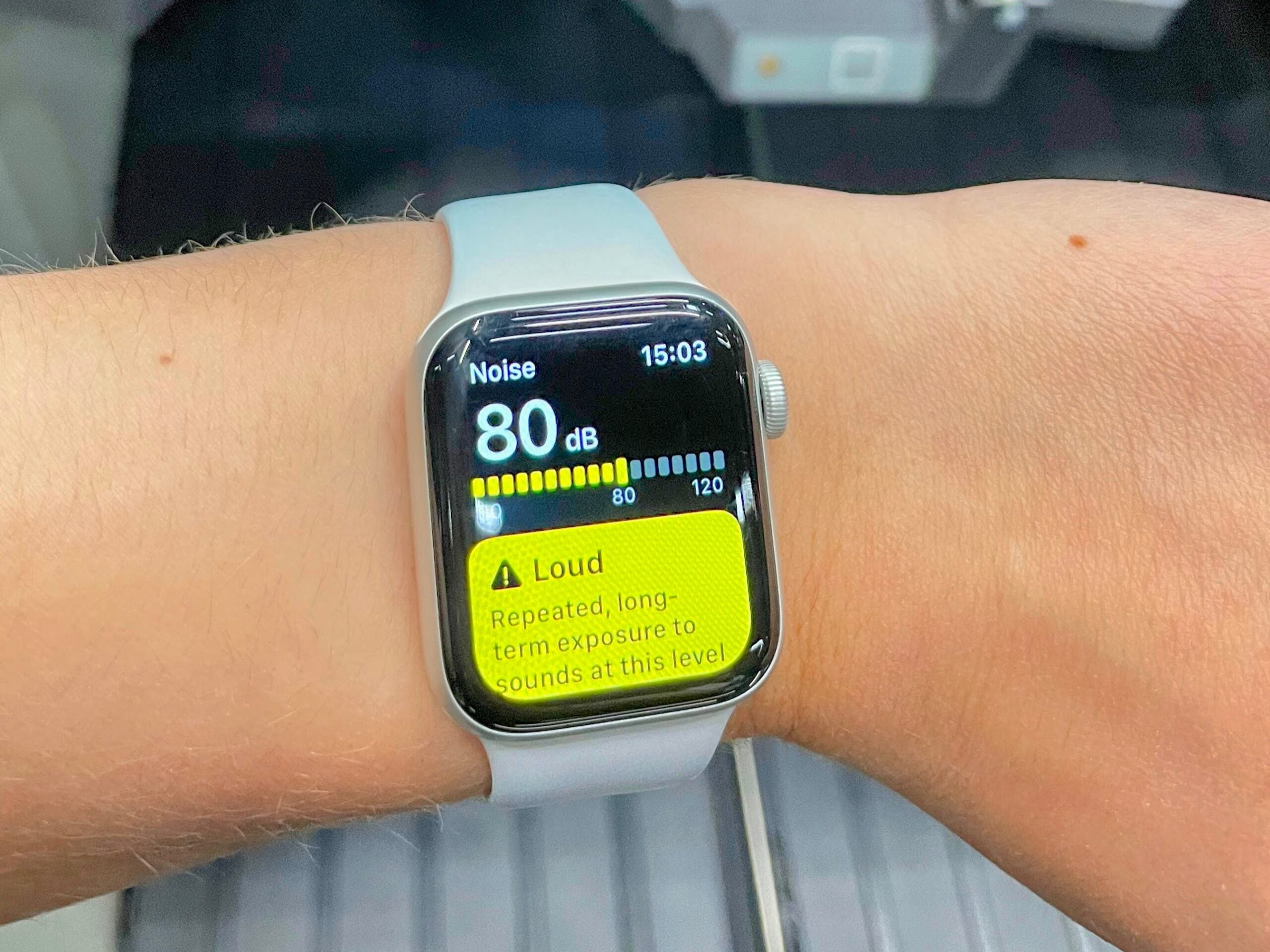 A smartwatch on a worker’s wrist shows a noise reading of 80 decibels and a “Loud” warning, held above the ballot trays of a mail sorting machine.