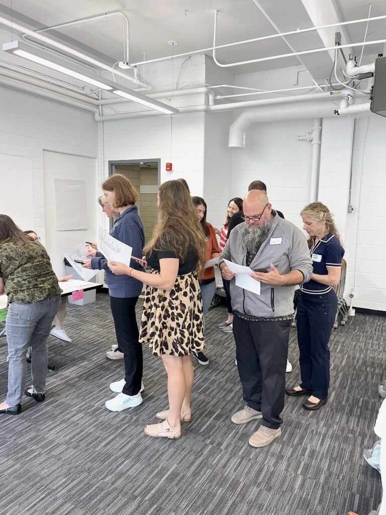 Adults wearing name tags and holding papers move between stations in a workshop room