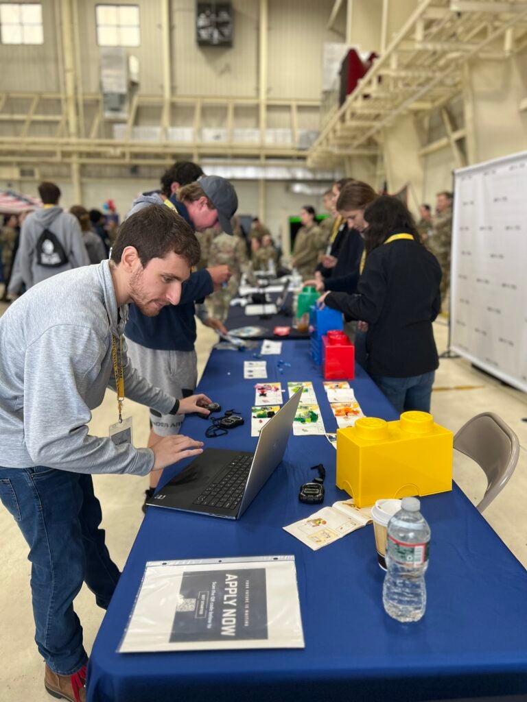 A young man leans over a laptop at a navy-draped table with LEGO bins and STEM activity sheets, people working behind him in a hangar