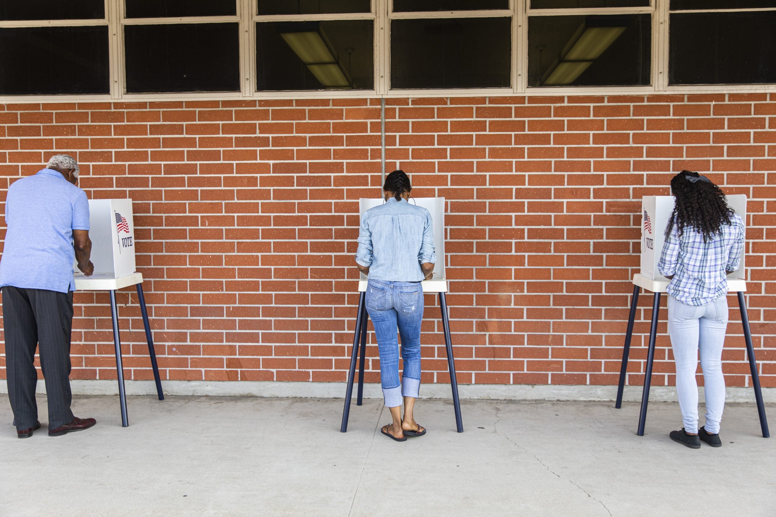 Voter seen from behind filling out a ballot at a privacy booth against a brick wall