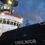 Ryan Cassin leans over the side of the research vessel Endeavor wearing a a blue sweatshirt with the letters URI