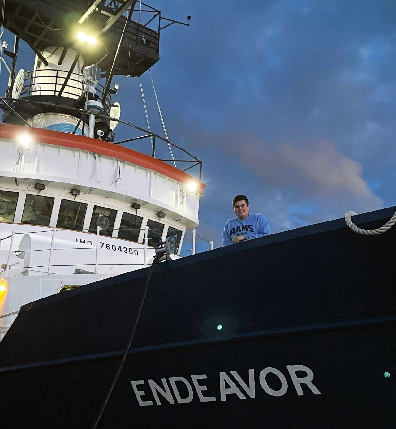 Ryan Cassin leans over the side of the research vessel Endeavor wearing a a blue sweatshirt with the letters URI