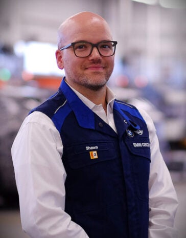 
Headshot of Shawn Drew wearing a navy blue BMW vest with branding and his name on it with a white collared shirt suit and checkered shirt and thick framed glasses with cars in the background.