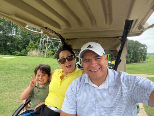Ana, her husband and her child take a selfie in a golf cart. All are wearing different colored polos, Ana is wearing sunglasses, her hsuband is wearing a white hat and her kid is making a peace sign.