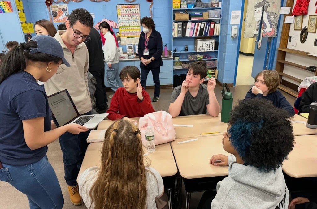 College students hold a laptop displaying language to a group of K-12 students