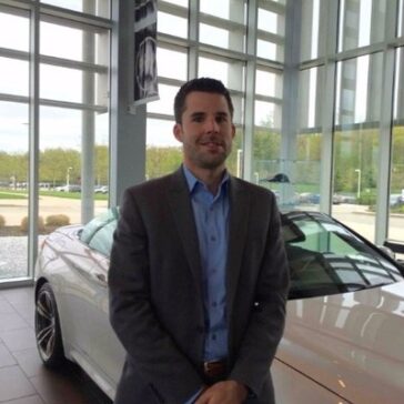 A photo of Eric Sargent wearing a dark suit and blue shirt in front of a wite car.