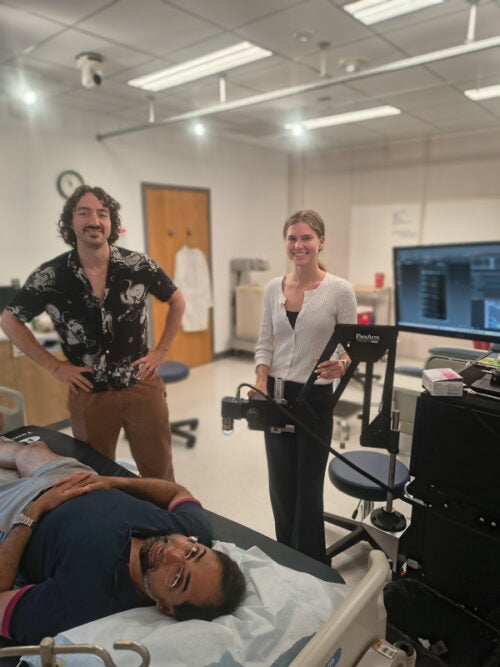 A photo of three researchers in a biomedical lab space, with Gabe and Heathre standing and Nevan on a hospital bed with technical instruments around him.