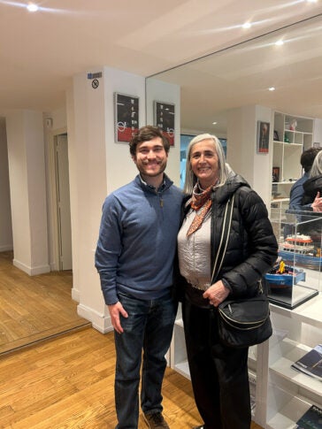Ian and Dr. Mattiauda are standing in a space with model boats on display smiling. Ian has a broad styled moustache, a blue jacket and jeans, while Dr. Mattiauda wears a black puffer jacket, a printed scarf and black pants.