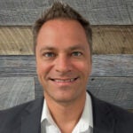 A professional headshot of Joel Karlsberg in front of a wood-textured background