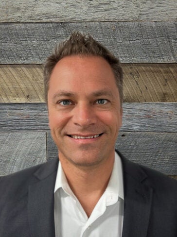 A professional headshot of Joel Karlsberg in front of a wood-textured background