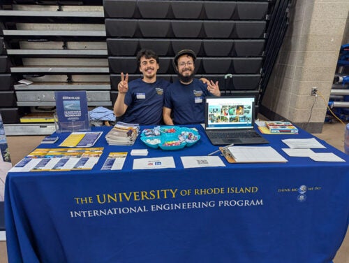 Jorge Menacho Aliaga and Jeremy Hererra Santos sit behind a table covered with a blue IEP tablecloth and several pamphlets and brochures about the IEP. Jorge is wearing a hat while Jeremy is making a peace symbol. Both are wearing IEP T-shirts.